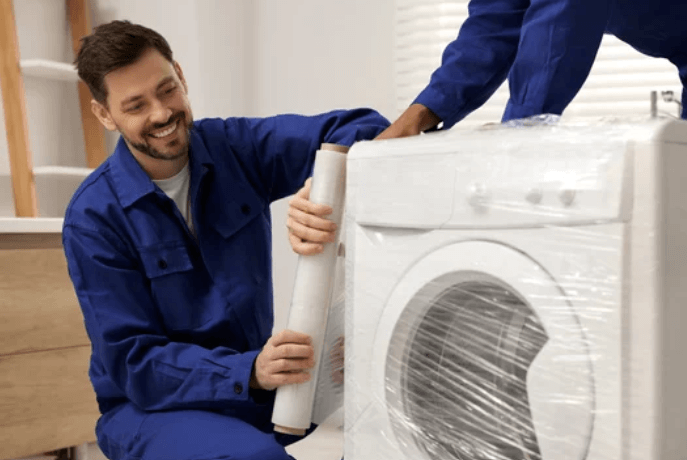 Un giovane uomo che, con la divisa blu, imballa con della pellicola una lavatrice, mentre il suo collega lo aiuta
