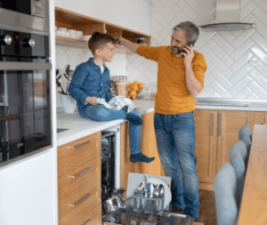 Un giovane bambino seduto sul piano lavoro della cucina mentre suo padre, affianco lui, fa una telefonata